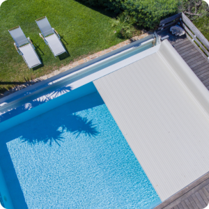 Aerial view of a blue rectangular pool with a white cover on a wooden deck, two lounge chairs on the nearby lawn.